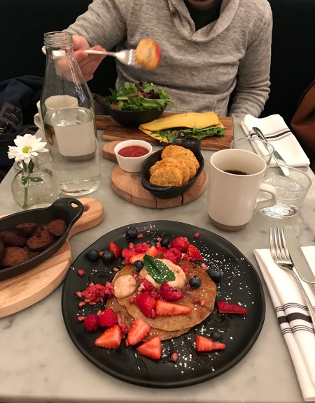 A white marble table piled with a plate of pancakes, a tureen of sliced sausages, a bowl of hash browns, and a plate with an omelette on it.
